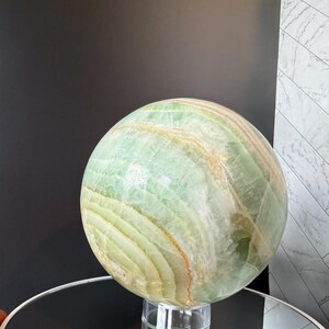 May include: A large, polished spherical stone featuring translucent layers of pale green, mint, and soft white with thin, warm amber-colored veins running throughout the surface. The sphere rests securely on a clear, circular acrylic display stand.