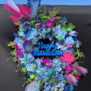 May include: A decorative wreath featuring blue flowers, green foliage, and pink accents. The center displays a blue sign with the text Happy Mardi Gras. It includes bright pink leaf-shaped cutouts, blue glittery sprigs, and green beaded strands.