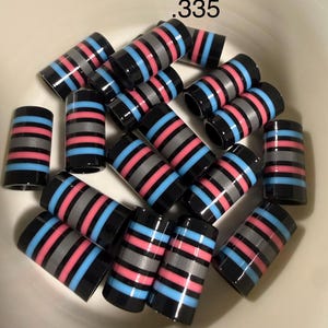May include: A collection of black, cylindrical beads with horizontal stripes in gray, pink, and blue. The beads are scattered in a white bowl. The text ".335" is in the upper right corner.