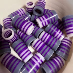 May include: A collection of cylindrical beads with a gray base and alternating purple and white stripes. The beads are scattered in a white bowl, showcasing their design and color variations. The beads are likely used for jewelry making or crafts.