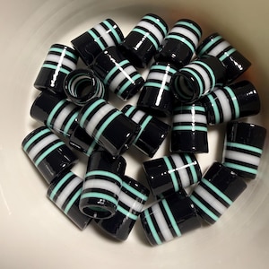 May include: A collection of black glass beads with horizontal stripes of white and light blue. The cylindrical beads are scattered in a white bowl, showcasing their glossy finish and contrasting colors.