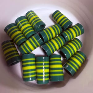 May include: A collection of cylindrical beads with a dark green base and horizontal yellow stripes. The beads are arranged in a white bowl, showcasing their vibrant color contrast and design.