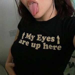 May include: Black crop top with the text "My Eyes are up here" in a white, bold font. The top is sleeveless and has a round neckline. The image shows a person with their tongue out.