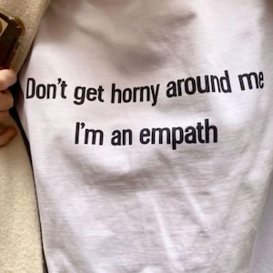 May include: White t-shirt with the black text "Don't get horny around me I'm an empath". The shirt is made of a soft material and is slightly wrinkled. The text is centered and easy to read.