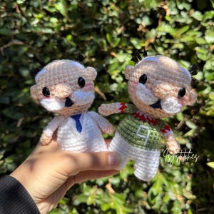 May include: Two small crochet dolls with light skin, black hair tufts, and white mustaches. One doll wears a white shirt with a blue tie, while the other wears a green jersey with a white number nine and red trim. Both have black eyes.