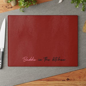 May include: Red cutting board with the text "Baddie in the kitchen" in black and pink lettering.