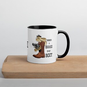 Könnte beinhalten: Weiße Keramik-Tasse mit schwarzem Innenraum und Henkel. Die Tasse zeigt eine Cartoon-Illustration einer Schlange mit Hut und Pistole in einem Cowboy-Stiefel. Der Text auf der Tasse lautet "There's a Snake in my Boot."