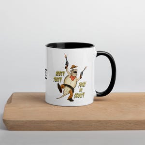 Può includere: Tazza in ceramica bianca con interno e manico neri. La tazza presenta un'illustrazione a cartoni animati di una lucertola con un cappello da cowboy e due pistole. Il testo sulla tazza recita "Rooty Tooty Point and Shooty."