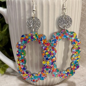 May include: A pair of silver hoop earrings with a colorful confetti design. The hoops are made of a scalloped edge and have a silver glitter circle at the top.