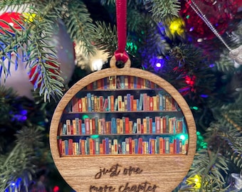May include: A round wooden Christmas ornament with a bookshelf design and the text "just one more chapter" engraved on the bottom. The bookshelf is filled with colorful books.