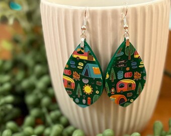 May include: A pair of green teardrop-shaped earrings with a camping theme. The earrings feature a pattern of colorful camper vans, tents, trees, and other camping-related imagery. The earrings are made of polymer clay and have silver hooks.