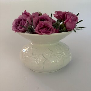 May include: A white ceramic vase with a bulbous base and a flared top, filled with pink roses and green thistle. The vase has a raised leaf pattern around the base. The arrangement is set against a white background.