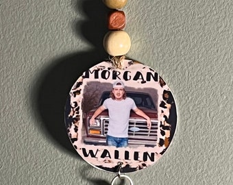 May include: A round keychain with a photo of Morgan Wallen in front of a truck. The keychain has a brown, tan, and white beaded strap and a silver clip.