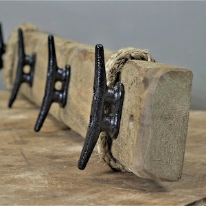 May include: A wooden coat rack with three black metal cleat hooks. The wood has a weathered appearance and a natural finish, with a rope detail. A nautical-themed home decor item.