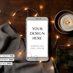 May include: A white smartphone mockup with a blank screen displaying the text "YOUR DESIGN HERE" and "SCREEN SIZE: 1080 x 1920 PX". The phone is on a wooden surface with a gray cloth, a lit candle, and string lights.