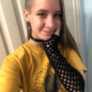 May include: A woman wearing a yellow t-shirt with a sunflower graphic and a black fishnet sleeve. She is wearing a black choker necklace and has short, shaved hair on one side.