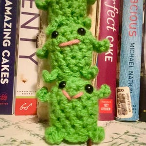 May include: A stack of three handmade green crochet frogs with black eyes and pink smiles. The frogs are positioned in front of a shelf of books, including "The Perfect Cake" and "Amazing Cakes."