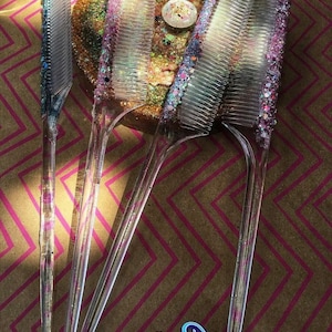 May include: Four clear plastic combs with glitter embedded in the plastic. The combs are arranged in a fan shape with the handles pointing down.