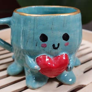 K&ouml;nnte beinhalten: Eine tealblaue Keramiktasse mit goldfarbenem Rand und einem niedlichen Comic-Gesicht. Die Tasse hat vier Beine und h&auml;lt ein rotes Herz in ihren H&auml;nden.
