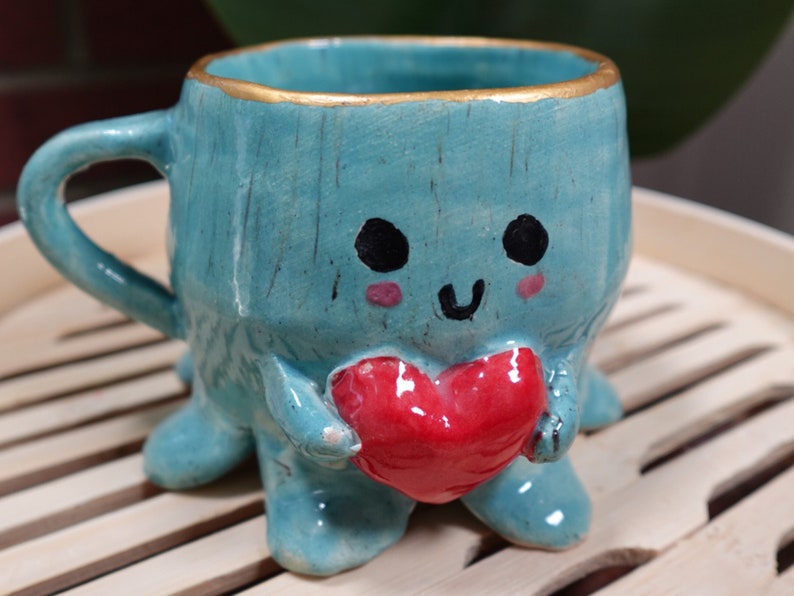 K&ouml;nnte beinhalten: Eine tealblaue Keramiktasse mit goldfarbenem Rand und einem niedlichen Comic-Gesicht. Die Tasse hat vier Beine und h&auml;lt ein rotes Herz in ihren H&auml;nden.
