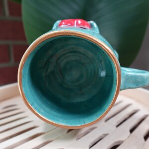 K&ouml;nnte beinhalten: Eine tealfarbene Keramiktasse mit goldfarbenem Rand und einem roten und wei&szlig;en Design auf der Oberseite. Die Tasse ist leer und das Innere ist sichtbar.