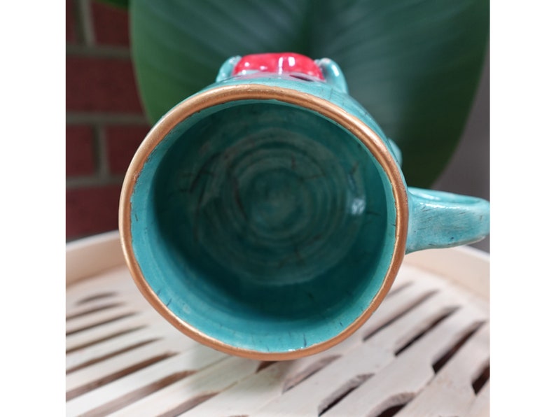 K&ouml;nnte beinhalten: Eine tealfarbene Keramiktasse mit goldfarbenem Rand und einem roten und wei&szlig;en Design auf der Oberseite. Die Tasse ist leer und das Innere ist sichtbar.