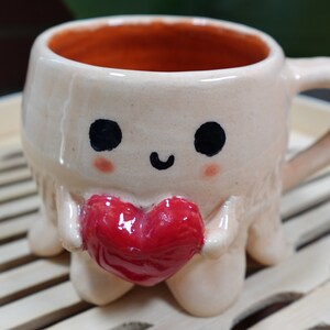 K&ouml;nnte beinhalten: Eine Keramiktasse in Form eines Oktopus mit einem roten Herzen in seinen Armen. Die Tasse hat eine wei&szlig;e Glasur mit schwarzen Augen und einem l&auml;chelnden Mund.