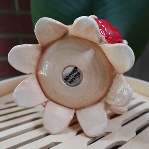 K&ouml;nnte beinhalten: Ein Keramik-Blumentopf mit einem roten und wei&szlig;en Blumendesign. Der Topf hat einen runden Boden und acht Bl&uuml;tenbl&auml;tter. Der Boden des Topfes hat einen schwarzen Stempel mit dem Wort "Happy" darauf.