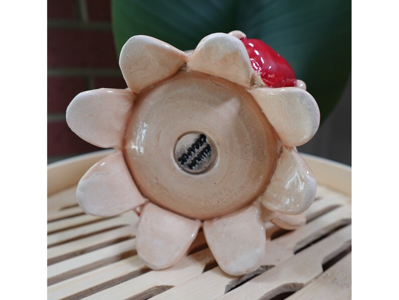 K&ouml;nnte beinhalten: Ein Keramik-Blumentopf mit einem roten und wei&szlig;en Blumendesign. Der Topf hat einen runden Boden und acht Bl&uuml;tenbl&auml;tter. Der Boden des Topfes hat einen schwarzen Stempel mit dem Wort "Happy" darauf.