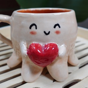 K&ouml;nnte beinhalten: Eine Keramiktasse in Form eines Oktopus mit einem l&auml;chelnden Gesicht und einem roten Herzen in der Hand. Die Tasse steht auf einer Holzoberfl&auml;che.