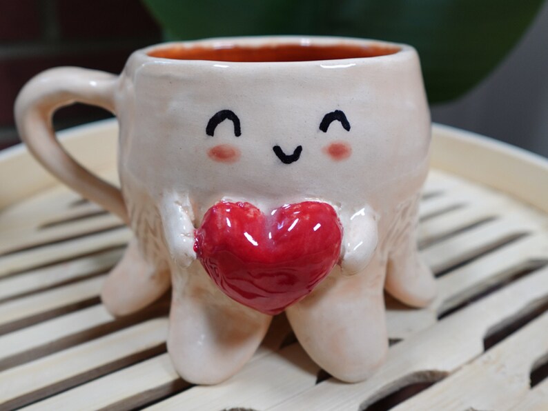 K&ouml;nnte beinhalten: Eine Keramiktasse in Form eines Oktopus mit einem l&auml;chelnden Gesicht und einem roten Herzen in der Hand. Die Tasse steht auf einer Holzoberfl&auml;che.