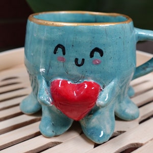 K&ouml;nnte beinhalten: Eine tealblaue Keramiktasse in Form eines Kraken, der ein rotes Herz h&auml;lt. Die Tasse hat einen goldenen Rand und ein handgemaltes Gesicht mit einem fr&ouml;hlichen Ausdruck.