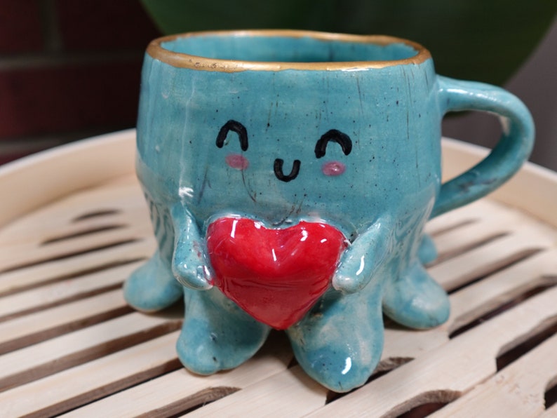 K&ouml;nnte beinhalten: Eine tealblaue Keramiktasse in Form eines Kraken, der ein rotes Herz h&auml;lt. Die Tasse hat einen goldenen Rand und ein handgemaltes Gesicht mit einem fr&ouml;hlichen Ausdruck.