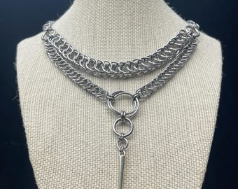 May include: A silver-colored chainmail choker necklace with a layered design. The necklace features two rows of chainmail, connected by a series of rings, and a dangling, pointed pendant. The jewelry is displayed on a mannequin.