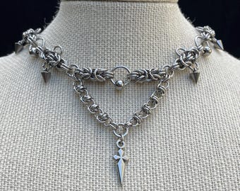 May include: A silver-toned necklace with a gothic aesthetic. The necklace features a chainmail-style design with a central pendant shaped like a dagger. It includes small cone-shaped spikes and decorative elements.