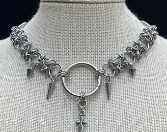 May include: A silver-toned chainmail choker necklace featuring a central circular ring, four hanging cone-shaped spikes, and a small cross pendant suspended from the bottom of the ring. The necklace is constructed from interlocking metal rings.