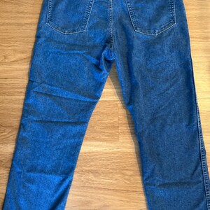 May include: Blue denim jeans with a classic five-pocket design. The jeans feature a button and zipper closure, belt loops, and the Wrangler brand patch on the back. The jeans are laid flat on a wooden surface.