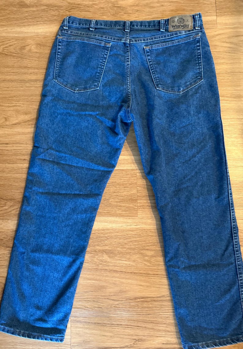 May include: Blue denim jeans with a classic five-pocket design. The jeans feature a button and zipper closure, belt loops, and the Wrangler brand patch on the back. The jeans are laid flat on a wooden surface.