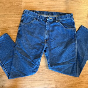 May include: Blue denim jeans with a classic five-pocket design. The jeans are laid flat on a wooden surface, showcasing the front and back pockets, belt loops, and button closure. The denim has a medium wash.