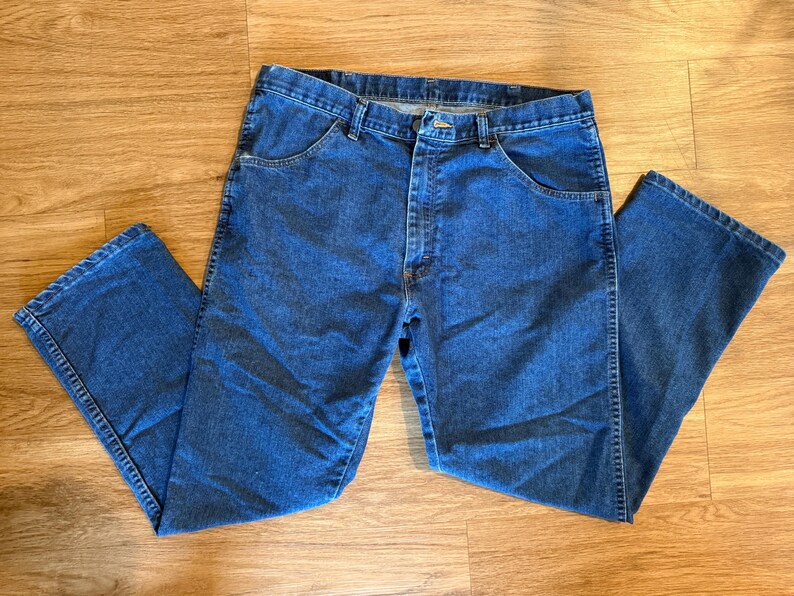May include: Blue denim jeans with a classic five-pocket design. The jeans are laid flat on a wooden surface, showcasing the front and back pockets, belt loops, and button closure. The denim has a medium wash.