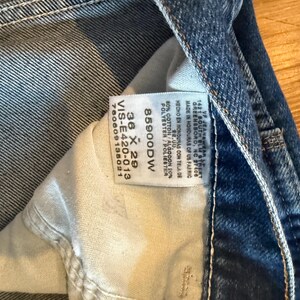 May include: Close-up of blue denim jeans with a white tag visible. The tag displays the size 36 x 29, along with the style number 85900DW and fabric composition. The jeans are made in Honduras of US fabric.