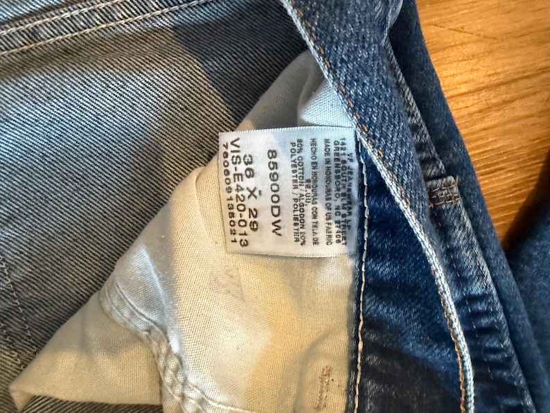 May include: Close-up of blue denim jeans with a white tag visible. The tag displays the size 36 x 29, along with the style number 85900DW and fabric composition. The jeans are made in Honduras of US fabric.