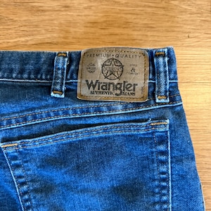 May include: Close-up of blue denim jeans with a brown leather patch. The patch features the Wrangler Authentic Jeans logo and text that reads "Premium Quality, Value, Authentic, Style & Comfort."