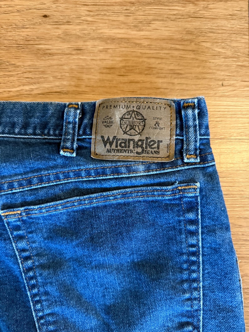 May include: Close-up of blue denim jeans with a brown leather patch. The patch features the Wrangler Authentic Jeans logo and text that reads "Premium Quality, Value, Authentic, Style & Comfort."