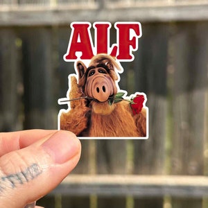 Könnte beinhalten: Ein Aufkleber mit ALF, einer pelzigen Alienfigur aus der Fernsehserie der 1980er Jahre, der eine rote Rose im Maul hält. Der Aufkleber trägt oben den Schriftzug "ALF" in roten Buchstaben.