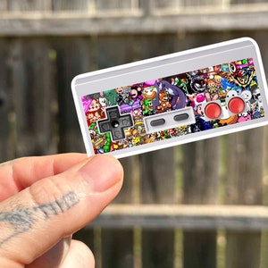 Może przedstawiać: Biała naklejka na kontroler Nintendo Entertainment System z kolorowym, pikselowym wzorem przedstawiającym różne postacie z gier wideo. Naklejka ma dwa czerwone przyciski i czarną krzyżówkę.