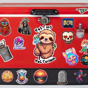 May include: A red metal cooler with various stickers, including a sloth holding a flower, a skull with hearts, a raccoon in a trash can, a Taco Bell logo, a skeleton with the text "Stay Positive", and a sunset landscape. The cooler has a black handle and a bottle opener.