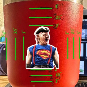 K&ouml;nnte beinhalten: Ein roter isolierter Becher mit einem wei&szlig;en Aufkleber eines Charakters, der ein rot-blaues Superman-Kost&uuml;m und einen rot-schwarzen karierten Hut tr&auml;gt. Der Becher ist mit Ma&szlig;en in Zoll gekennzeichnet: 2,5, 2, 2,5, 2, 2,5, 3, 2,5, 2, 2,5, 3.