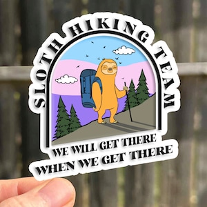 May include: A sticker with a cartoon sloth wearing a backpack and hiking with a stick. The sticker has a blue, pink, and purple background with clouds and trees. The text on the sticker reads "Sloth Hiking Team" and "We will get there when we get there."