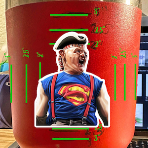 K&ouml;nnte beinhalten: Ein roter, rostfreier Becher mit einem wei&szlig;en Aufkleber eines Charakters aus dem Film 'Kevin - Allein zu Haus'. Der Charakter tr&auml;gt ein rot-blaues Hemd mit einem Superman-Logo, rote Hosentr&auml;ger und einen wei&szlig;en Piratenhut. Der Becher hat eine Messskala an der Seite mit Markierungen f&uuml;r 2,5 Zoll, 2 Zoll und 3 Zoll.